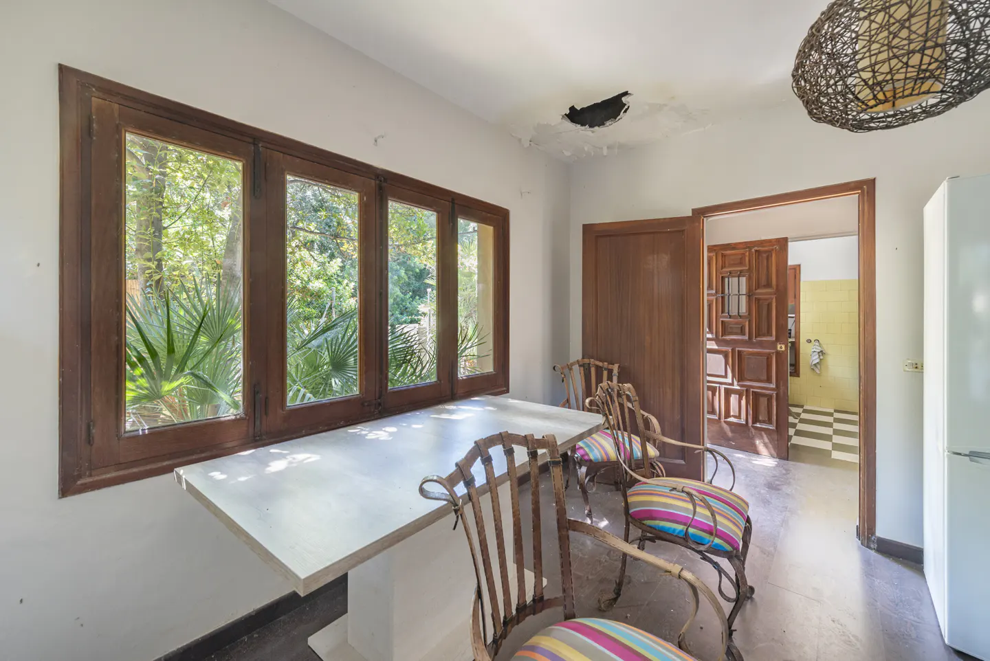Dining area with a white table, metal chairs with striped cushions, large wooden windows, and a damaged ceiling.