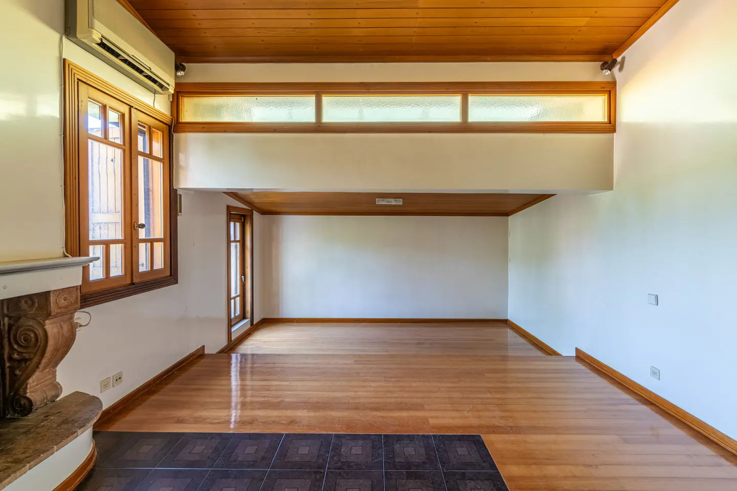 Empty room with hardwood floors, white walls, and a wood ceiling. A fireplace is on the left, and windows are on the back wall.