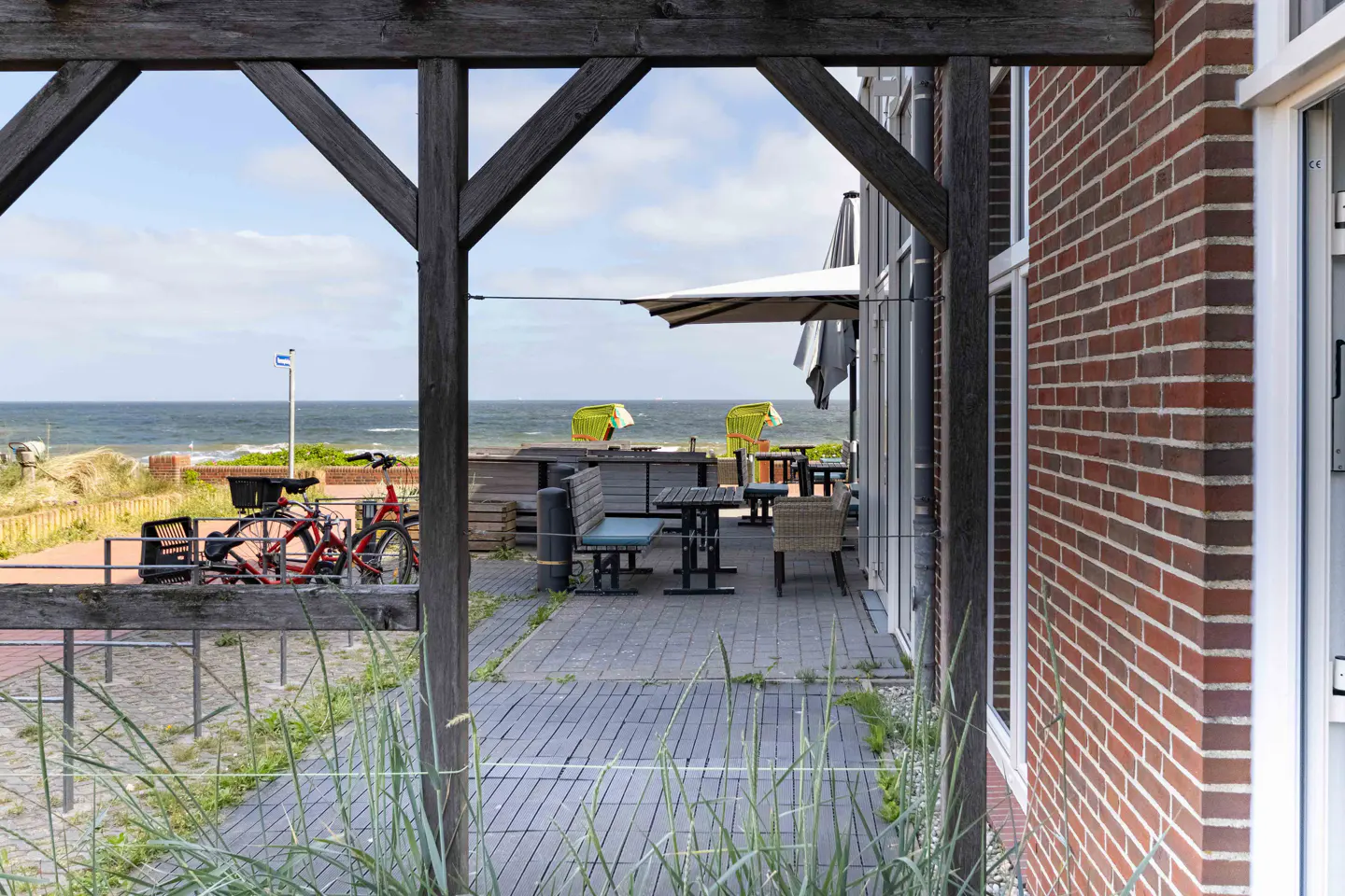 View of a seaside patio with outdoor seating, tables, and bicycles, framed by a wooden pergola and red brick building.