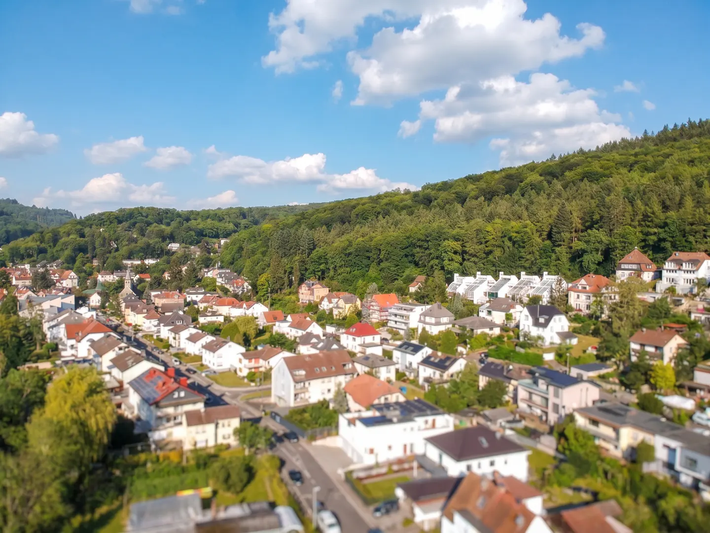 Aerial view of a European town with houses, roads, and a lush green forest under a blue sky with scattered white clouds.