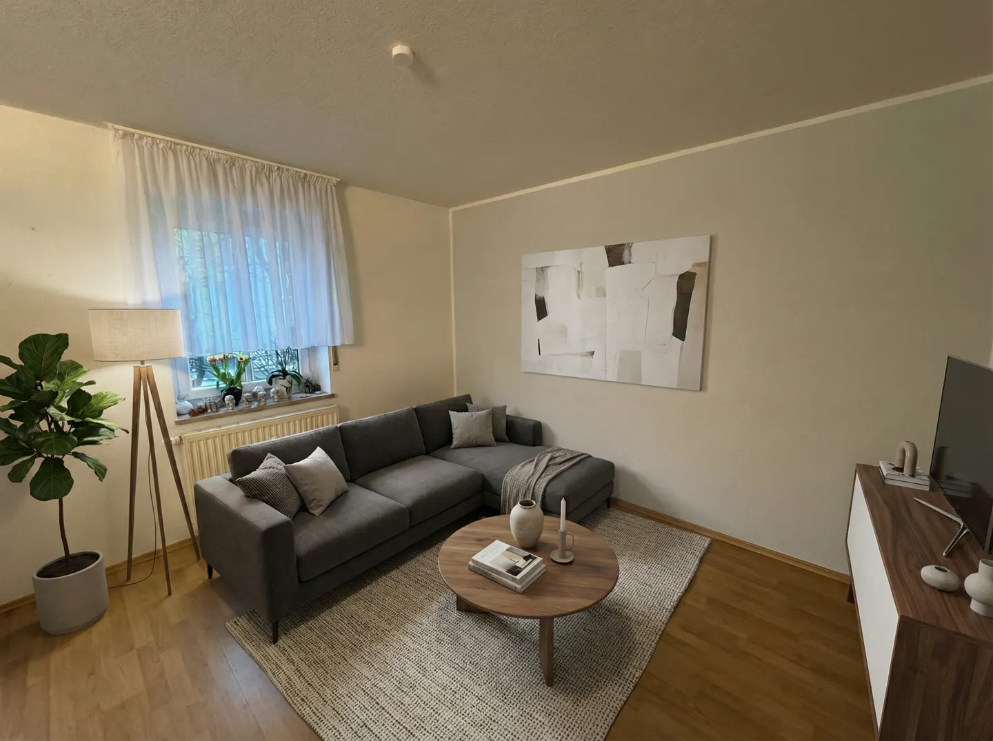 Living room with gray sectional sofa, round wood coffee table on a textured rug, and abstract art on the wall. A floor lamp and potted plant are near the window.