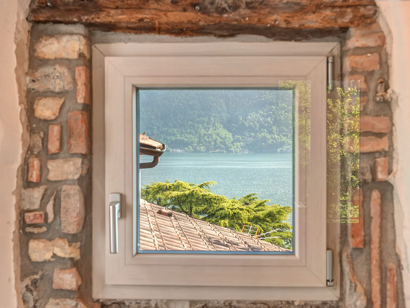 A window framed by brick and wood, overlooks a lake and green hills. A tiled roof and trees are visible below.