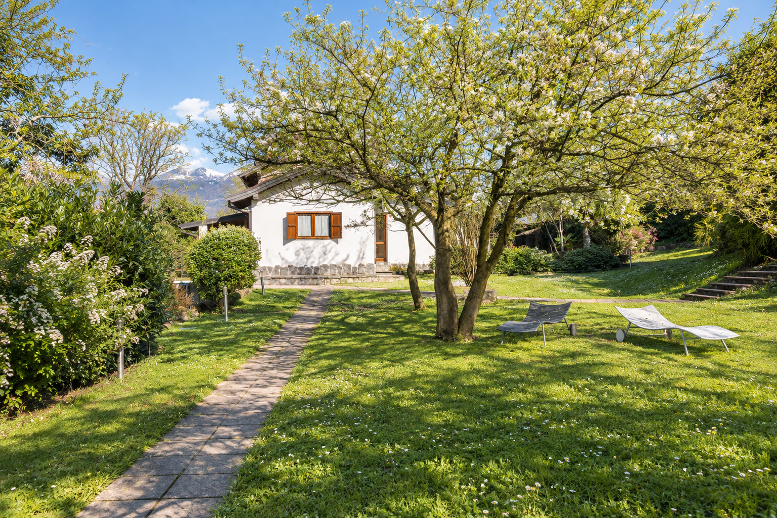White house with brown shutters in a lush green garden with a stone path, flowering trees, and two lounge chairs. [AI-generated text]