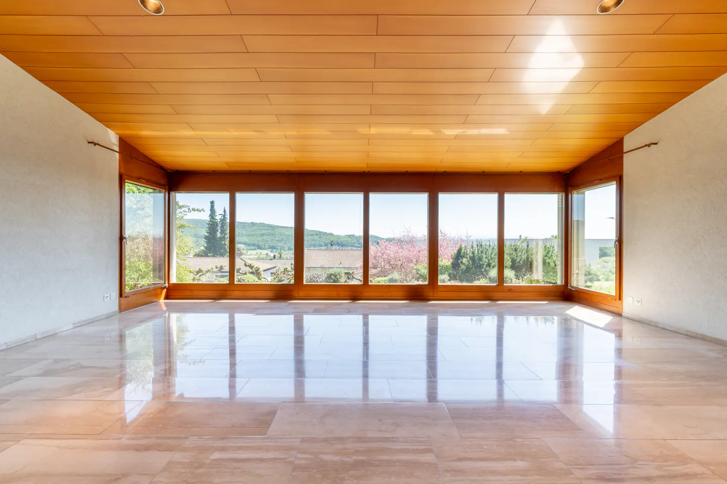 Empty room with wood ceiling, marble floor, and large windows. Outside view of trees and hills.