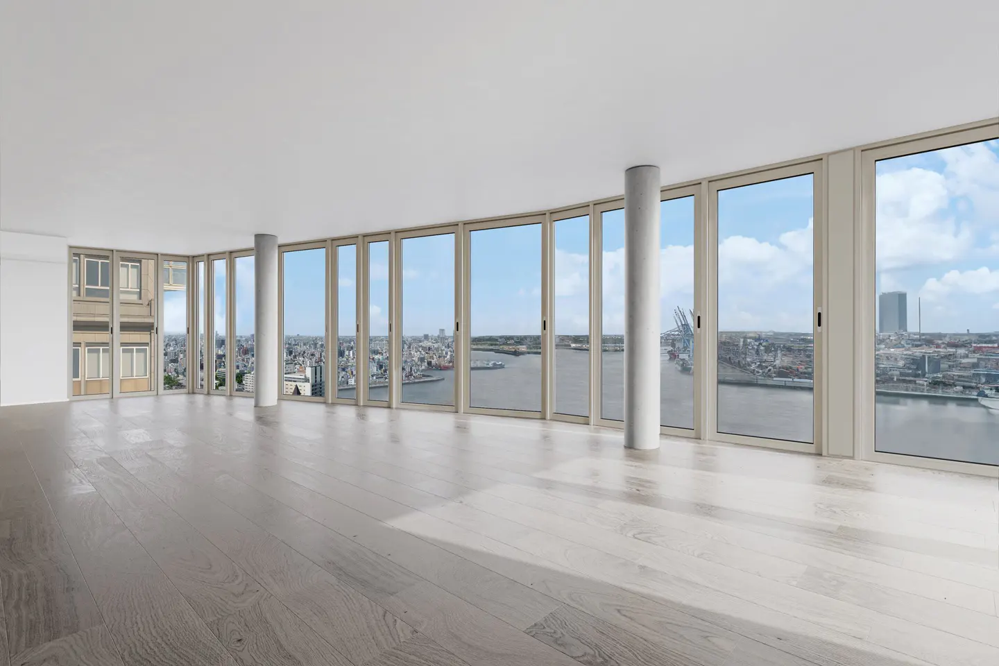 Empty room with light wood floors and white walls. Large windows offer a city view with water and blue sky.