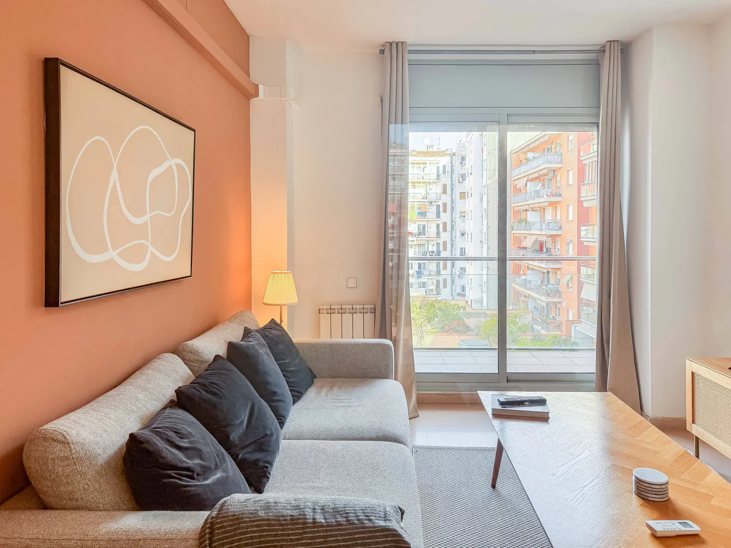 Living room with a gray sofa, black pillows, and a wooden coffee table. A large window shows city buildings. The wall is peach with abstract art.