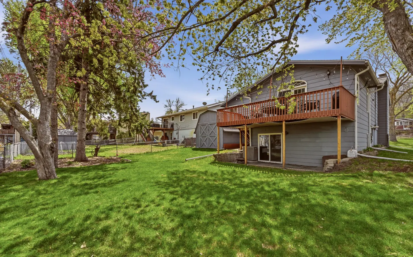 A gray two-story house with a brown deck overlooks a green lawn with trees and a blue sky.