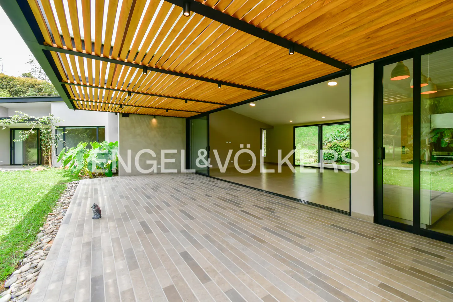 Exterior view of a modern home with a wood slat pergola, a stone patio, and large glass doors. The Engel & Völkers logo is visible.