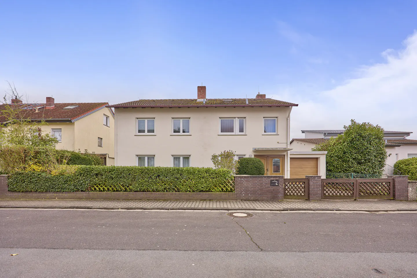 Two-story beige house with a brown roof and white-framed windows. A green hedge and a brown fence line the front yard. Blue sky above.