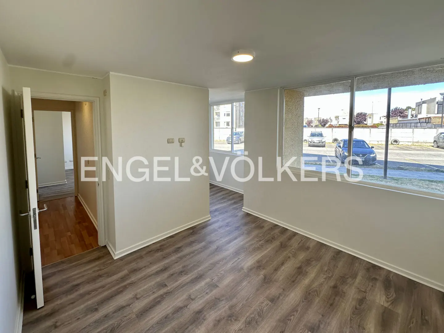 Bright, empty room with gray wood floors, white walls, and large windows overlooking a parking lot. Doorway to another room is visible.