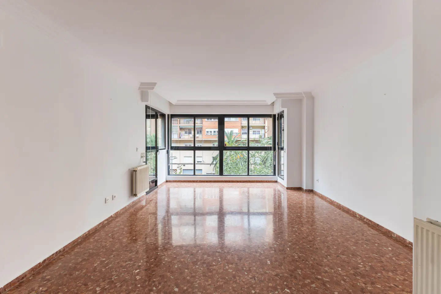 Empty room with white walls, a red speckled floor, and a large window with a view of trees and buildings.