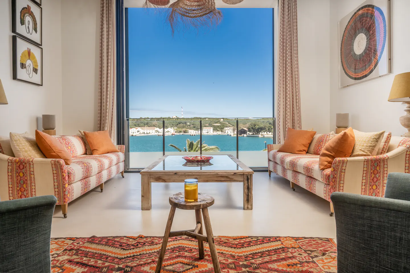 Bright living room with patterned sofas, wood table, and ocean view through a large window. An orange rug adds warmth.