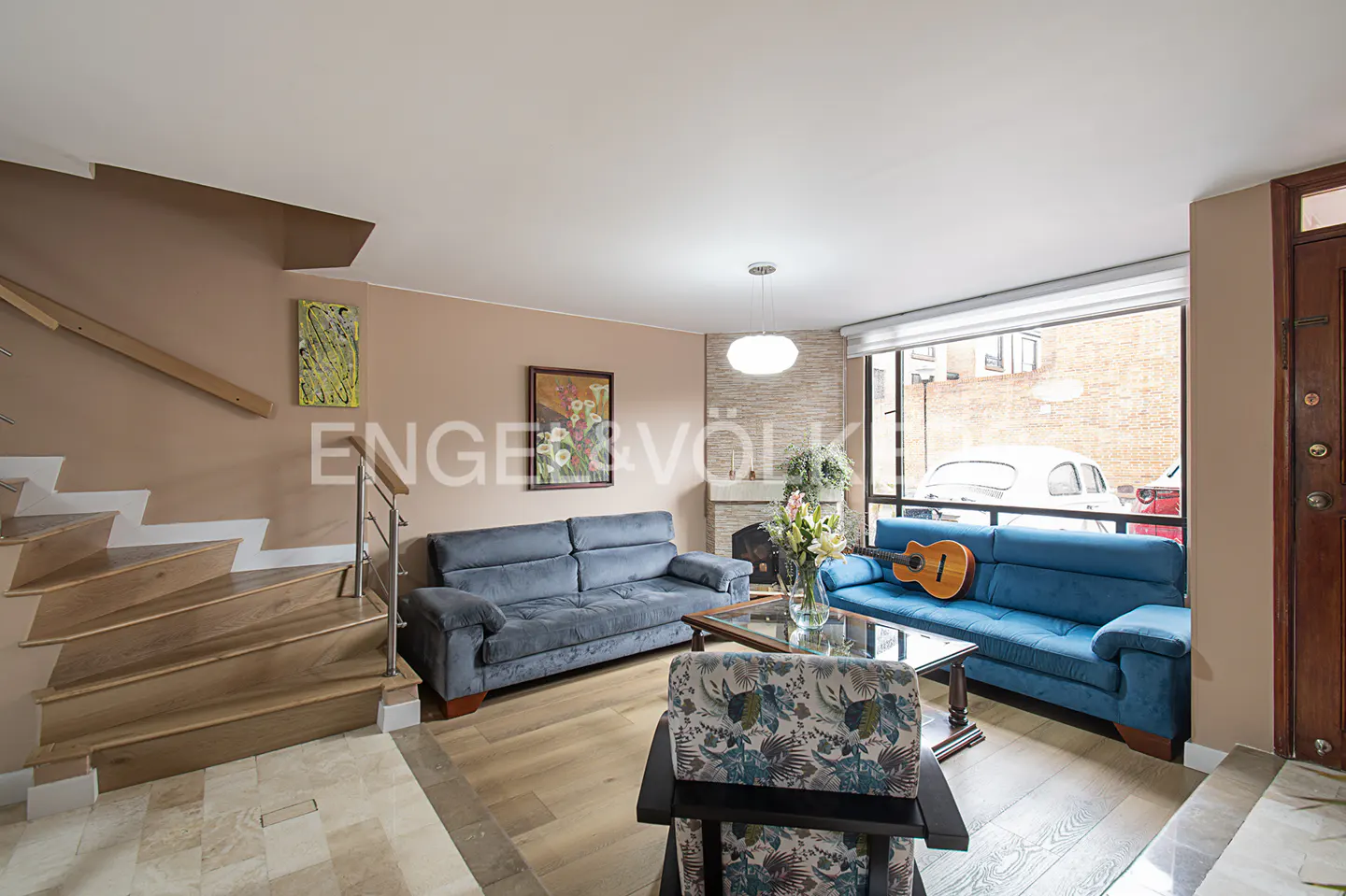 Living room with blue sofas, a glass coffee table, and a guitar on the sofa near a large window. Stairs are on the left.