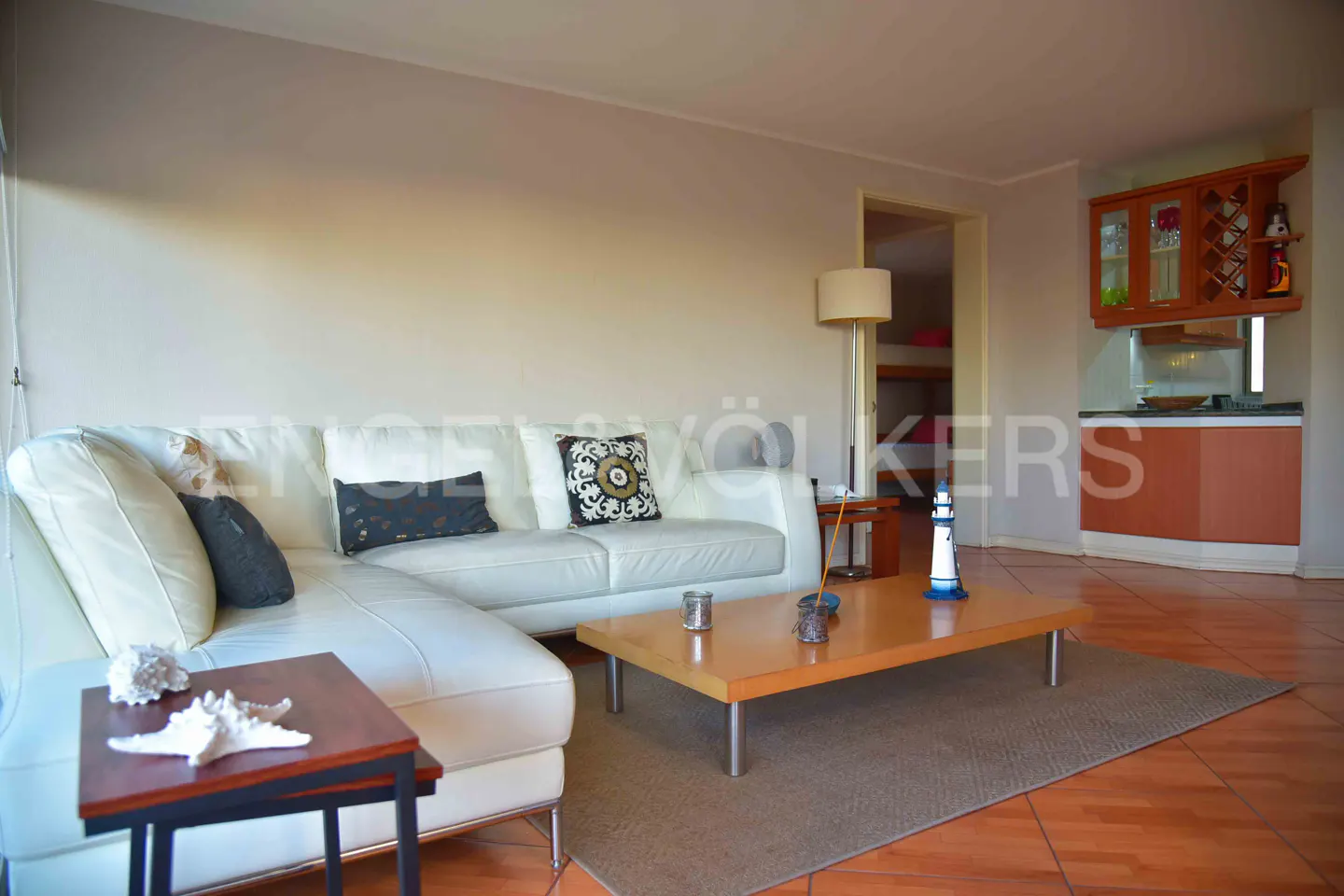 Living room with white leather sofa, wood coffee table, and nautical decor. Kitchenette in background.