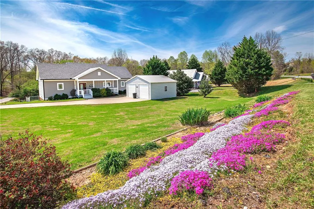 A gray house with a white porch and a gray shed sit on a green lawn with a flower bed.