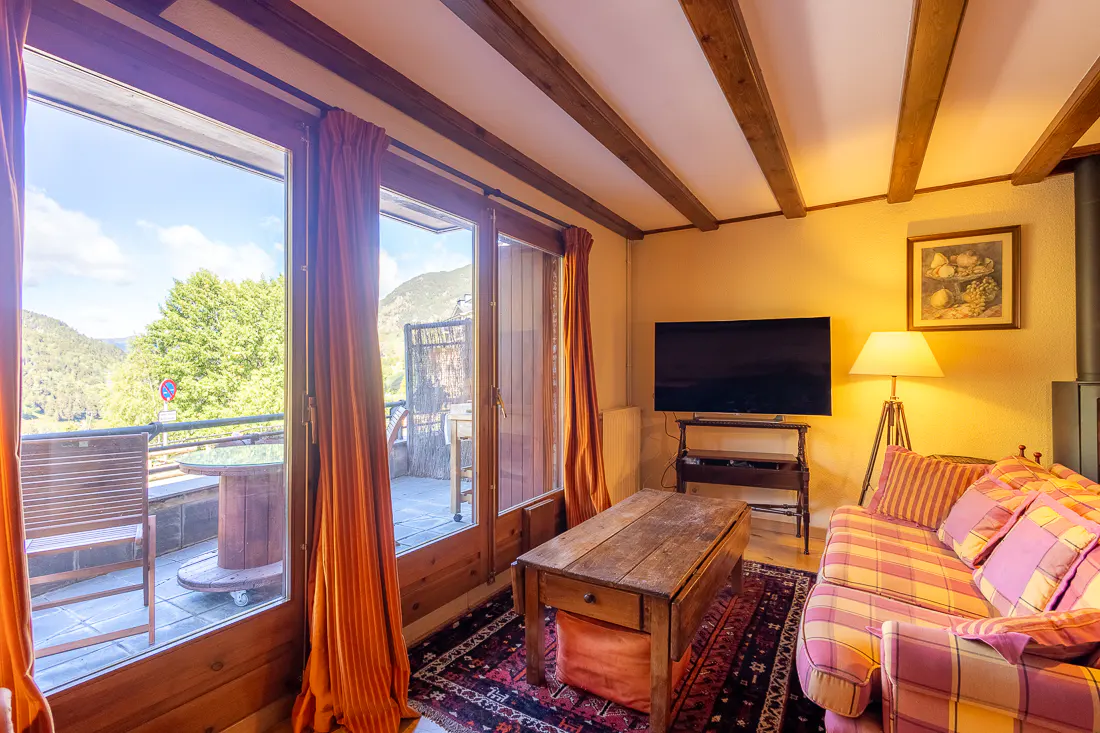 Cozy living room with a plaid sofa, wooden coffee table, TV, and large glass doors opening to a mountain view balcony.