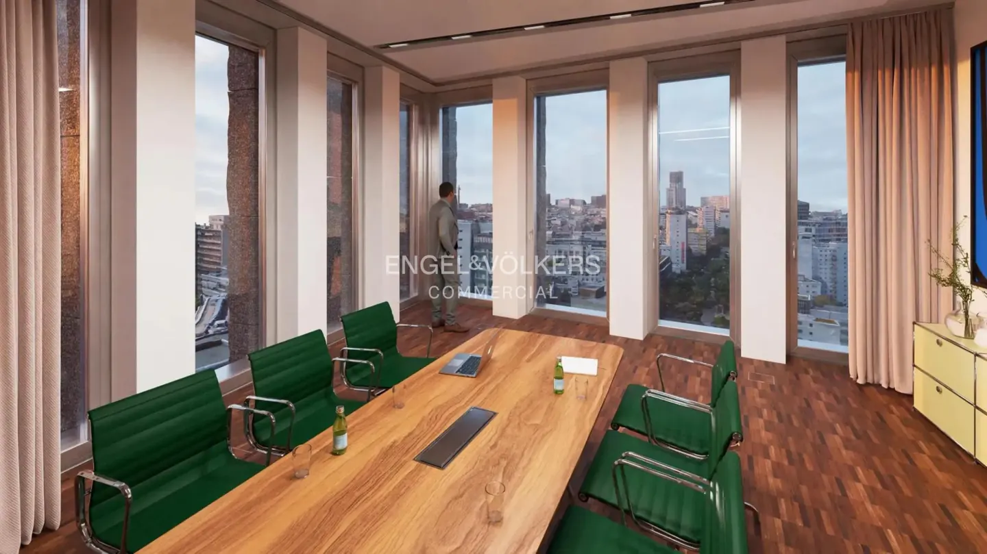 Modern office boardroom with a wooden table, green chairs, and a man looking out floor-to-ceiling windows at a city view.