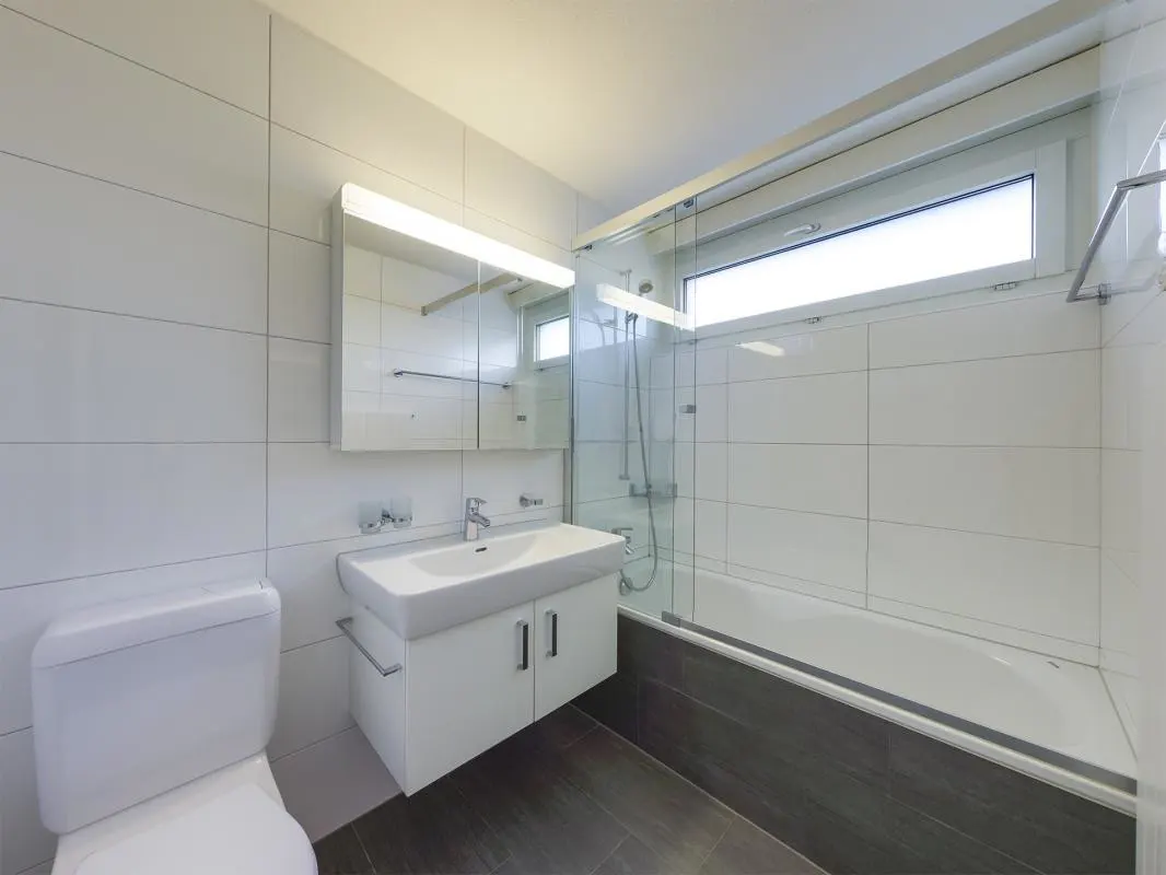 Modern bathroom with white tiled walls, floating vanity, toilet, and a bathtub with a glass shower screen.