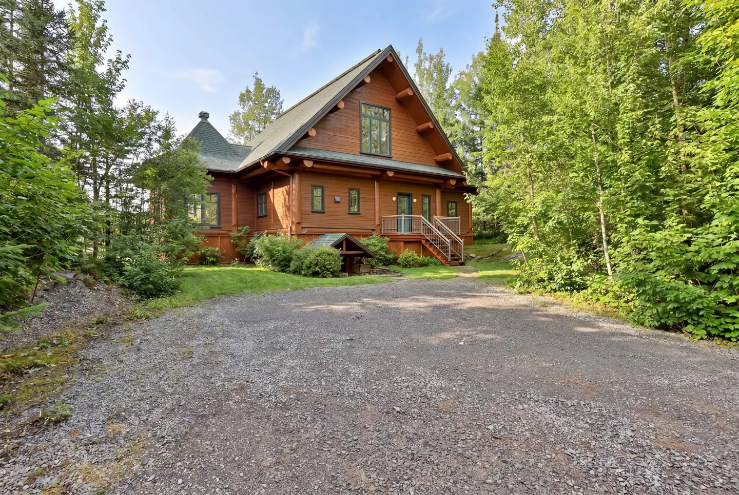 A two-story log cabin with a green roof, surrounded by trees and a gravel driveway.