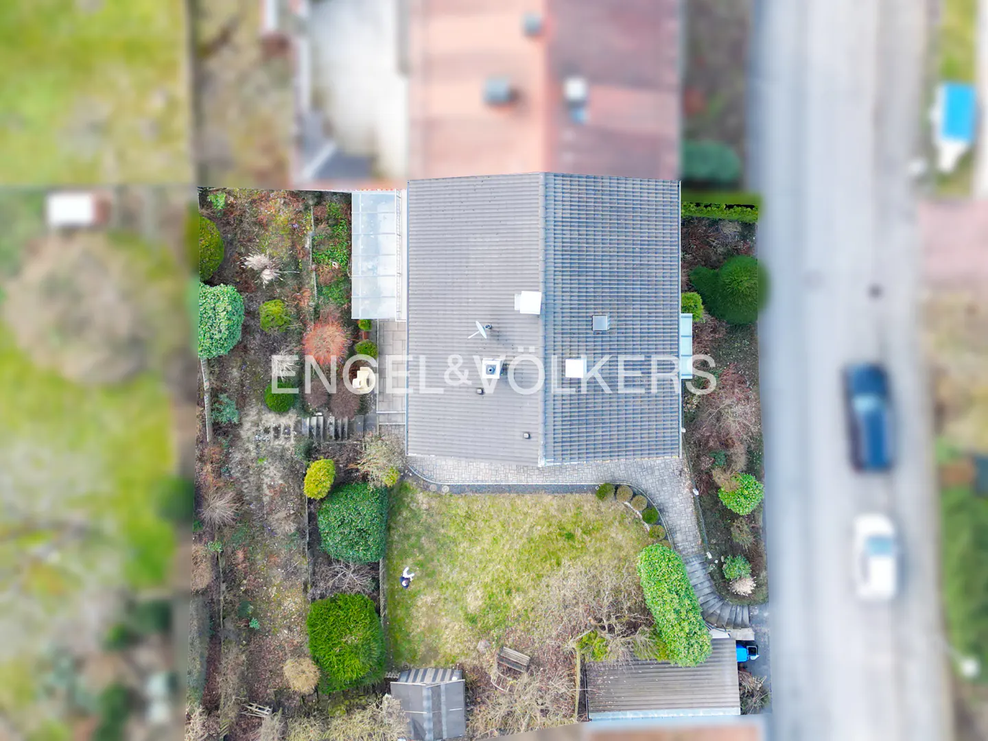 Aerial view of a house with a gray roof, green lawn, and garden, with the Engel & Völkers logo. A road with cars is visible in the background.