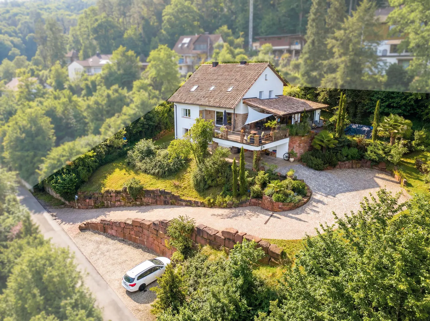 Aerial view of a white two-story house with a brown roof, surrounded by lush greenery and a stone wall. A white car is parked in the driveway.