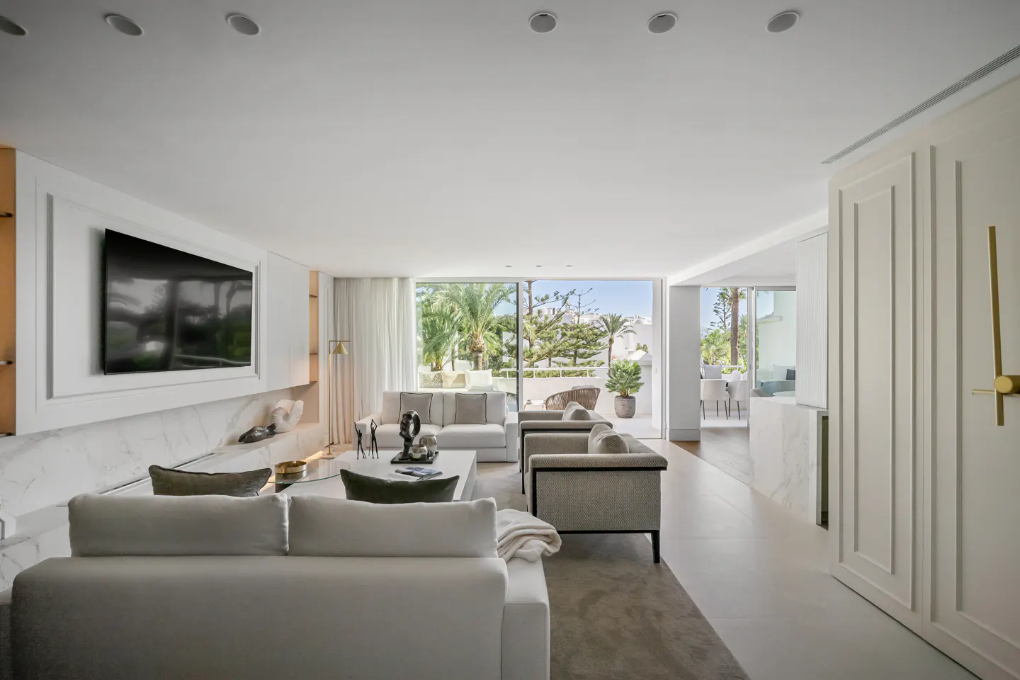 Bright, modern living room with white sofas, chairs, and marble accents. Large windows offer a view of palm trees and a sunny patio.