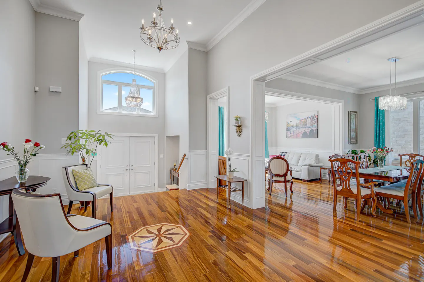 Bright foyer with hardwood floors, white walls, and high ceilings. A dining room is visible through an archway. Chandeliers hang from the ceiling.