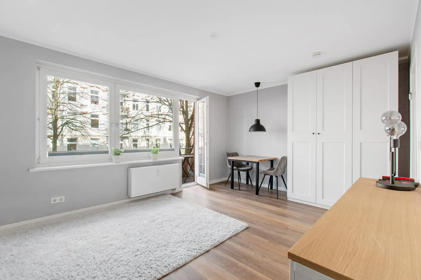 Bright apartment interior with wood floors, white rug, and gray walls. A table and chairs sit near an open balcony door.
