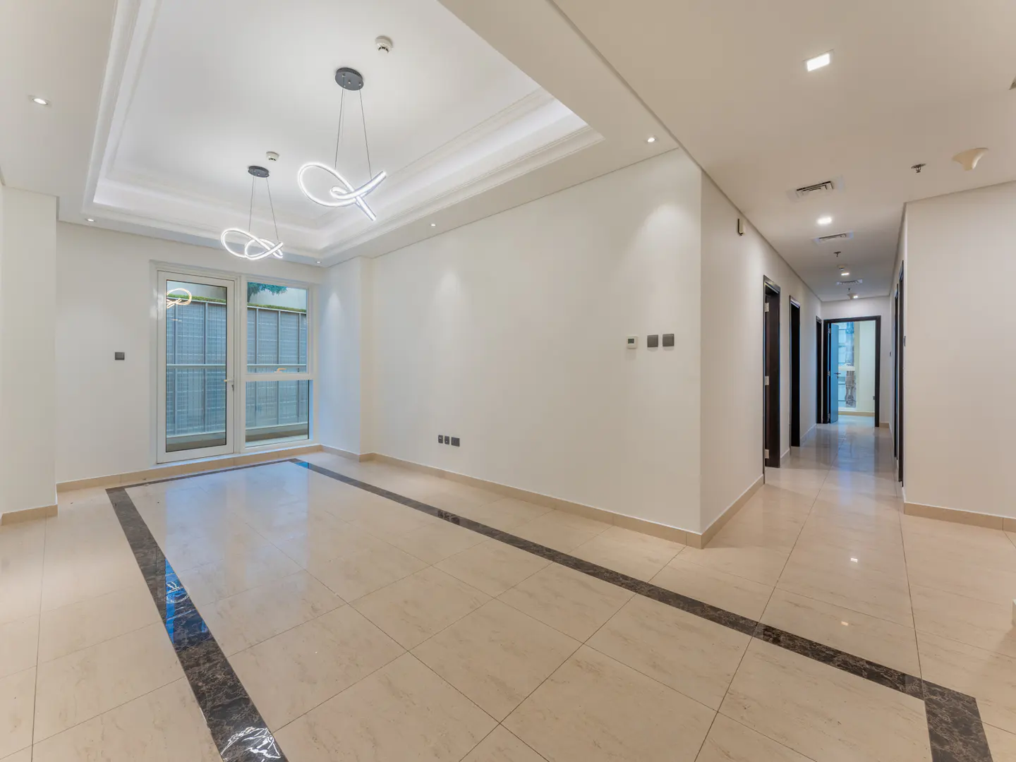 Bright, empty room with beige tile floor, white walls, and modern light fixtures. A hallway with dark doors extends to the right.