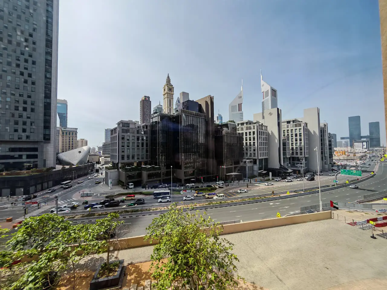 Cityscape view of Dubai with modern buildings, a busy highway, and green trees in the foreground.