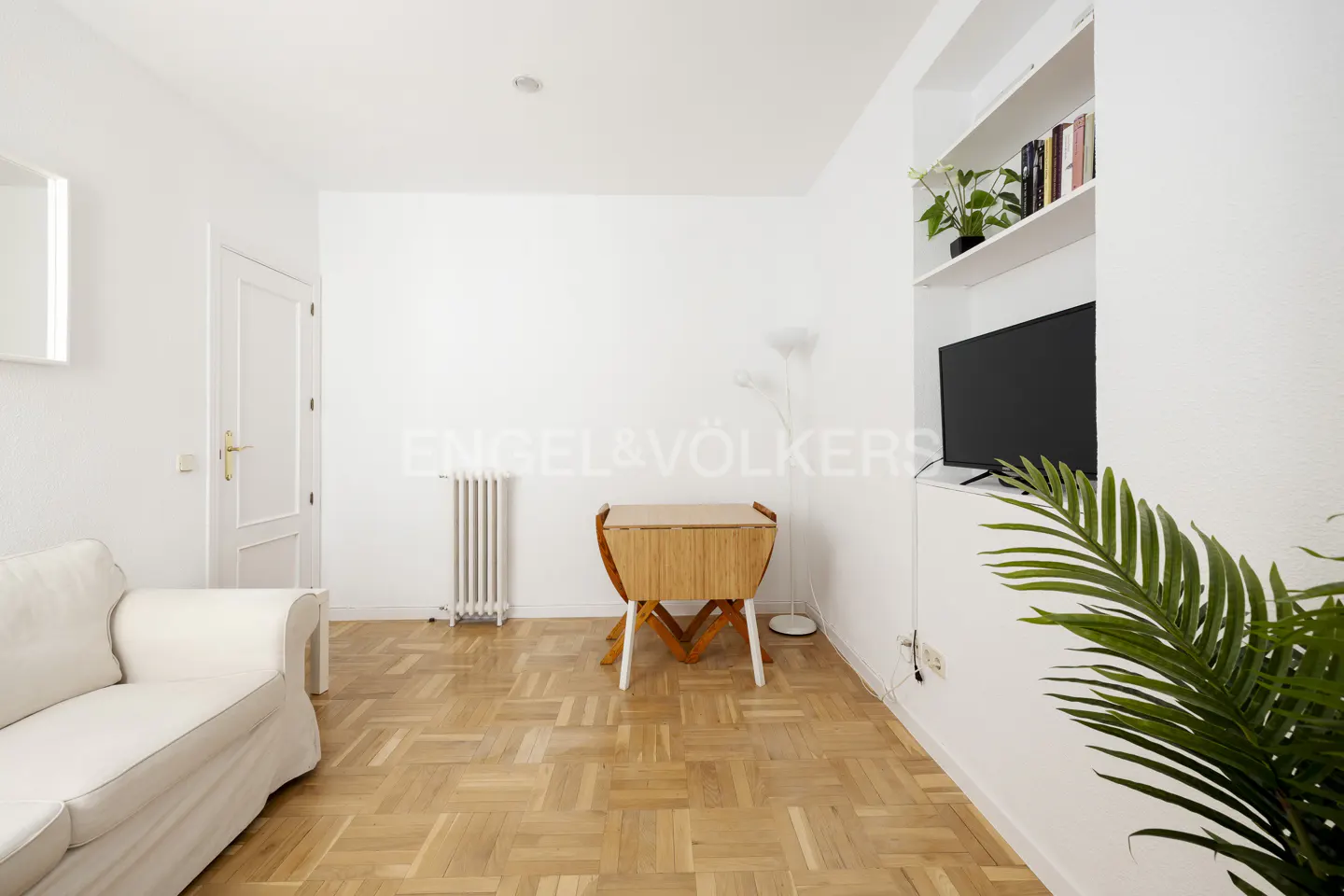 Bright living room with white walls, parquet floor, white sofa, radiator, folding table, TV, and built-in shelves.