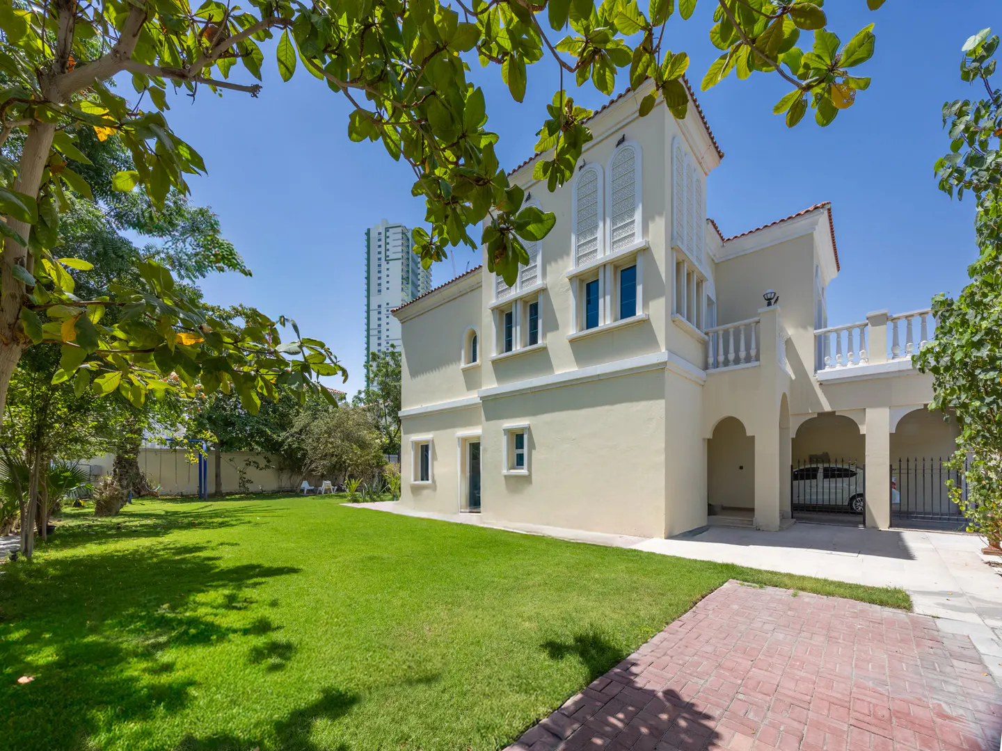 Two-story beige house with a red tile roof, green lawn, and brick walkway under a blue sky. A tall building is in the background.