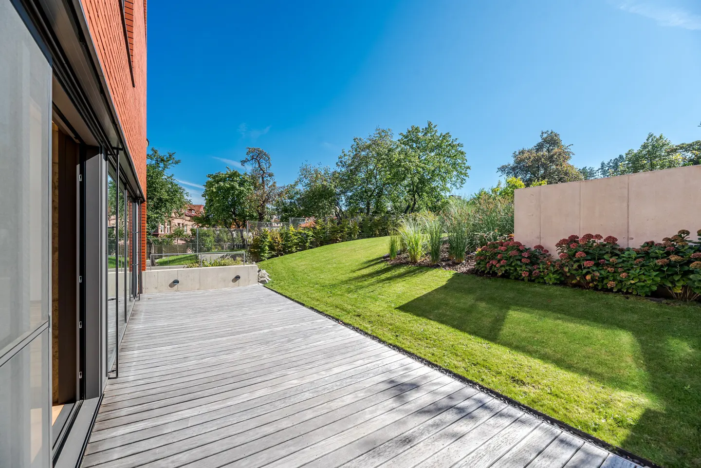 A modern home's backyard features a gray wood deck, green lawn, trees, and a clear blue sky. A brick wall and glass doors are visible.