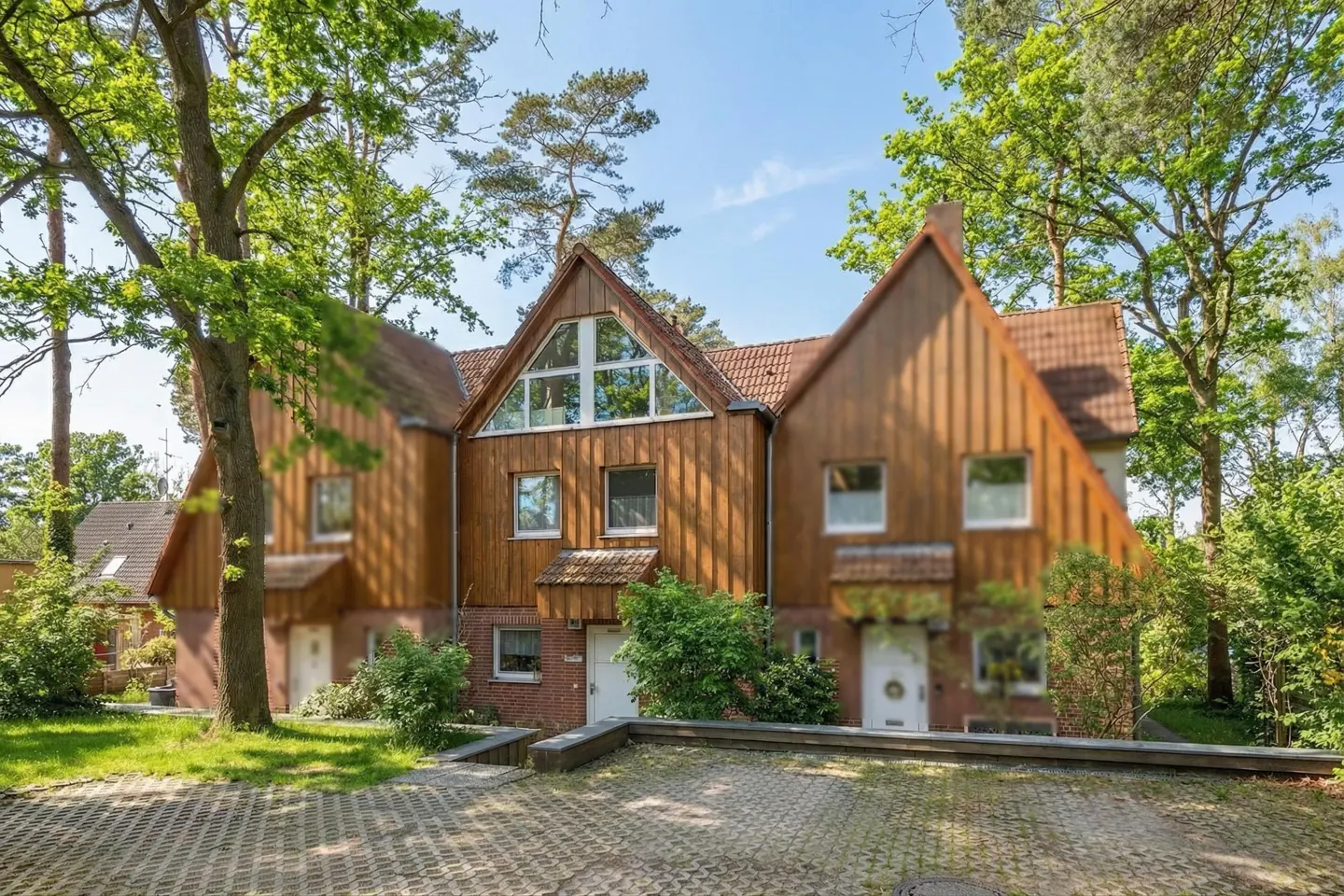 A three-unit townhouse with brown wood siding, red tile roof, white doors and windows, and a gray brick driveway. Trees surround the building.