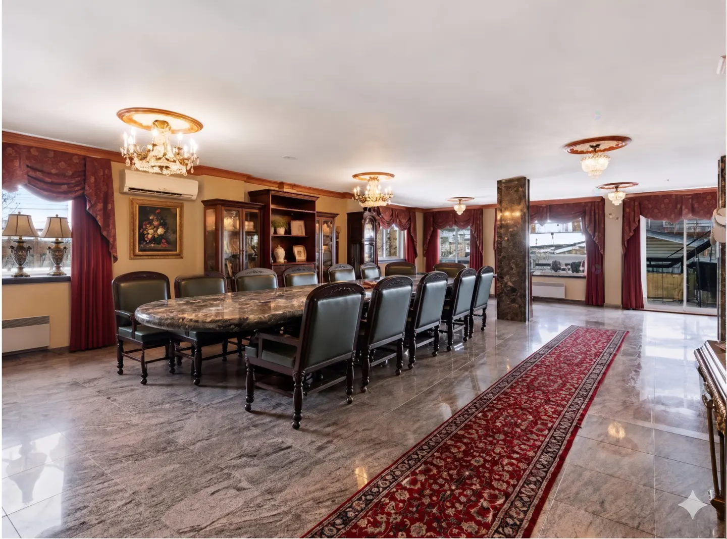A spacious dining room with a long marble table, leather chairs, and a red patterned rug. Chandeliers hang from the ceiling.