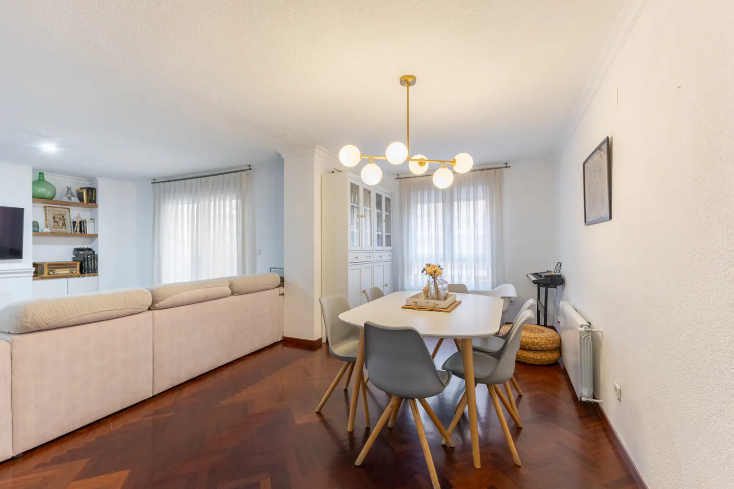 Bright living and dining area with a long beige sofa, white walls, and dark wood floors. A white dining table with gray chairs sits under a modern gold chandelier.