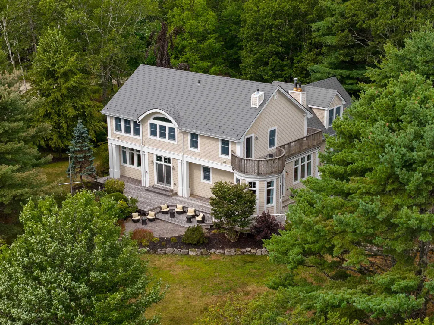 Aerial view of a beige two-story house with a gray roof, surrounded by green trees and a wooden deck with patio furniture.