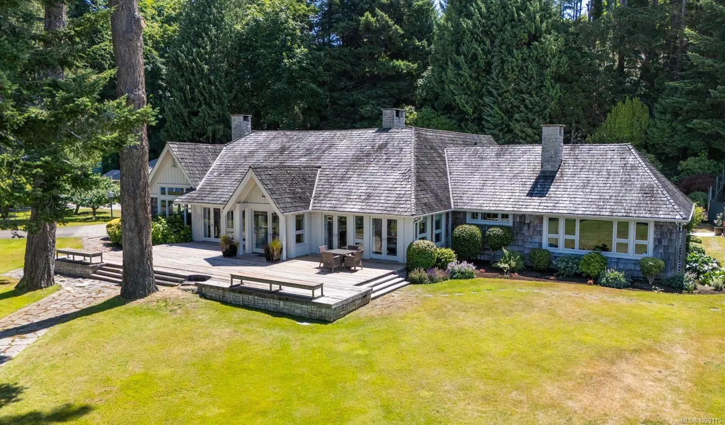 Aerial view of a white house with a gray shingle roof, surrounded by green trees and a lawn. A wooden deck with benches and a table extends from the house.
