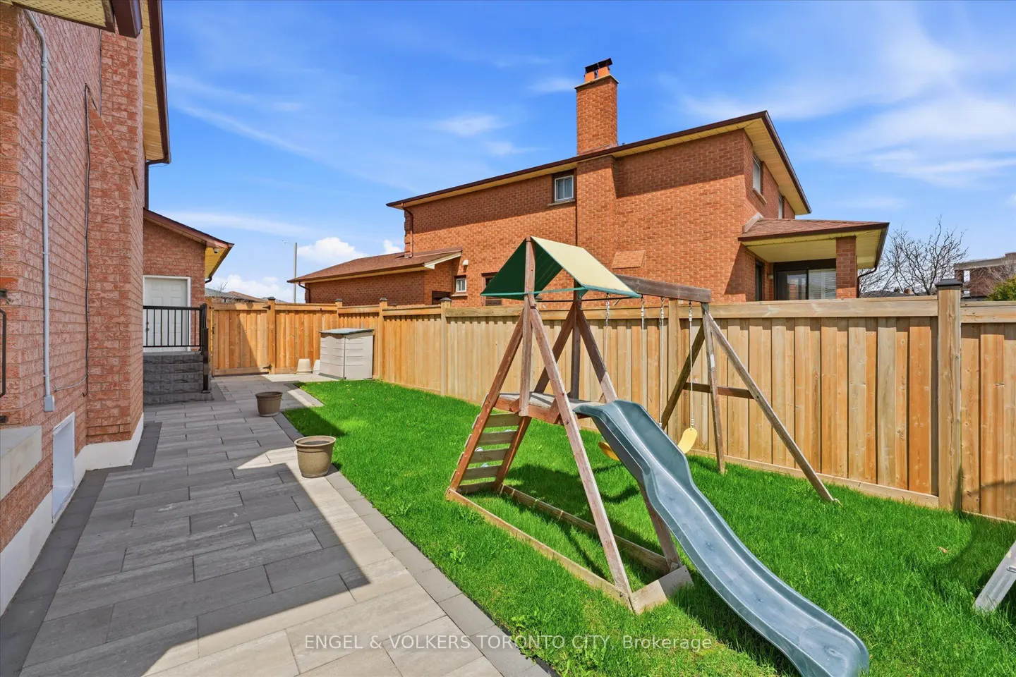 Backyard with a stone patio, green lawn, wooden fence, and a children's wooden playset with a slide under a blue sky.