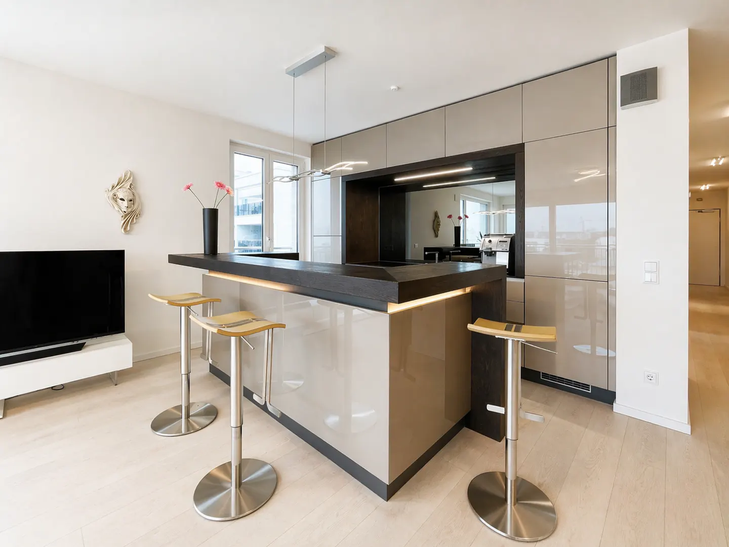 A modern kitchen with a dark countertop island, four metal stools, and beige cabinets. A TV and mask are on the left wall.