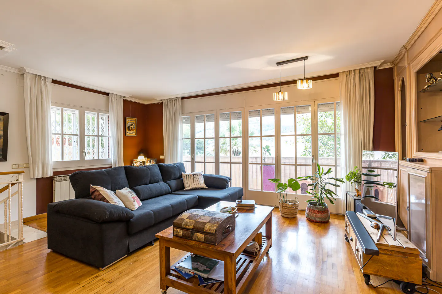 Bright living room with a blue sectional sofa, wooden coffee table, and large windows with white frames and curtains. Hardwood floors and plants add warmth.
