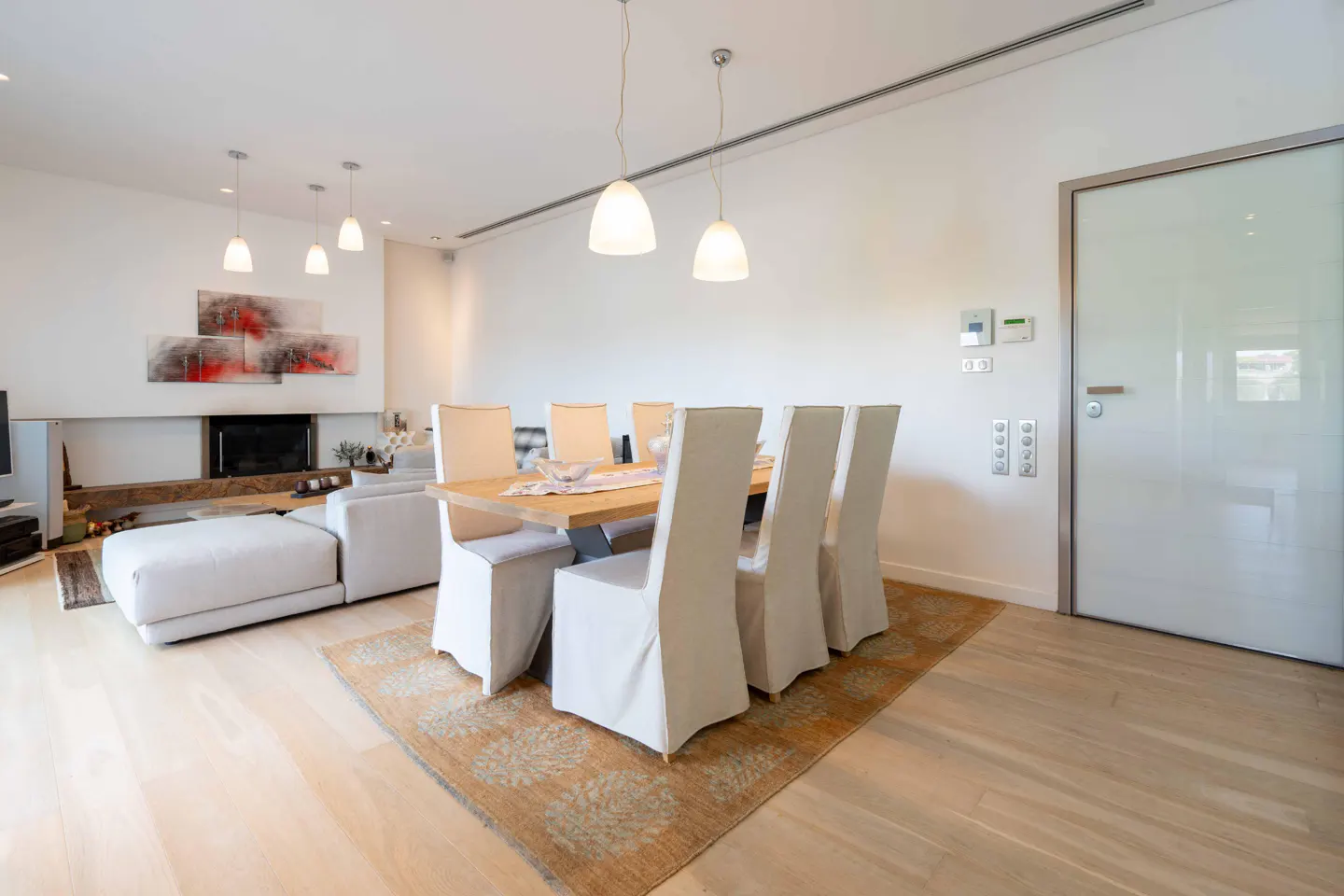 Bright, modern dining area with a light wood table, white chairs, and a patterned rug on a light wood floor. Living room in the background.