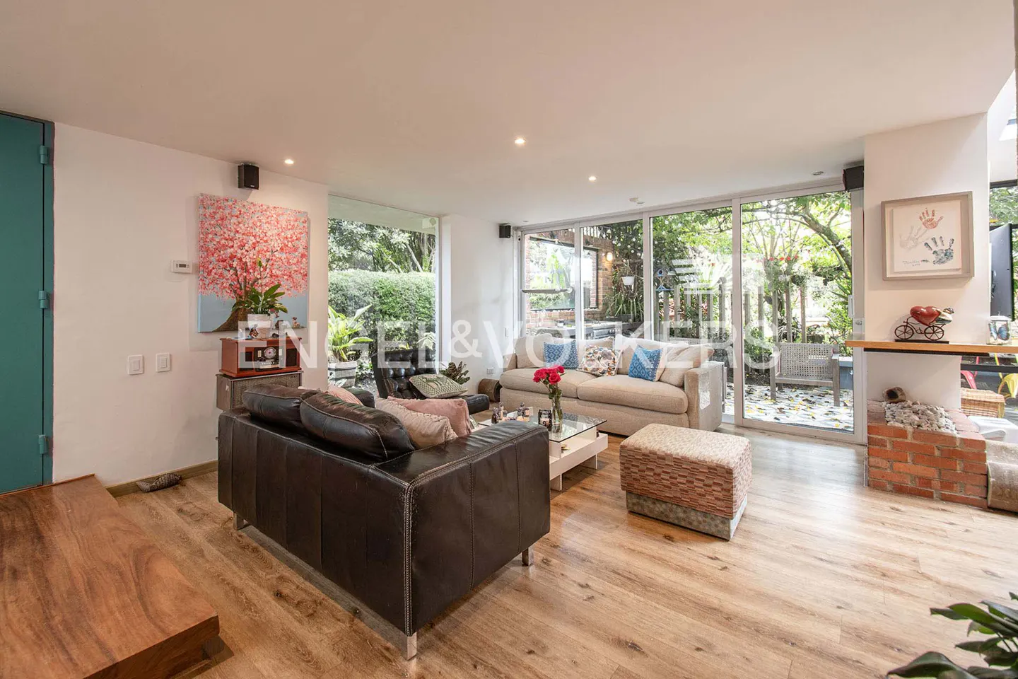 Living room with hardwood floors, a black leather sofa, and a beige sofa facing a glass wall with a garden view.