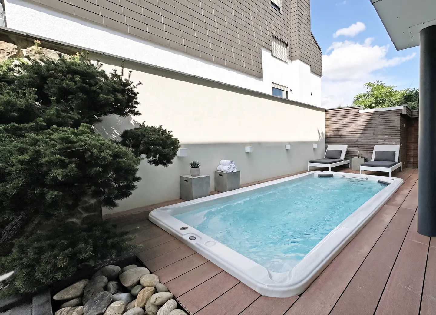 Outdoor pool with lounge chairs on a wood deck, next to a building with a white wall and a green bush.