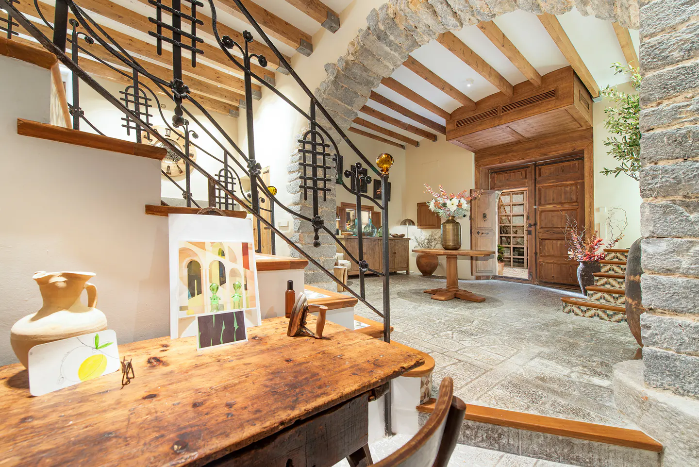 Interior view of a rustic home with stone walls, wooden beams, and a wrought iron staircase. A wooden desk with artwork sits in the foreground.