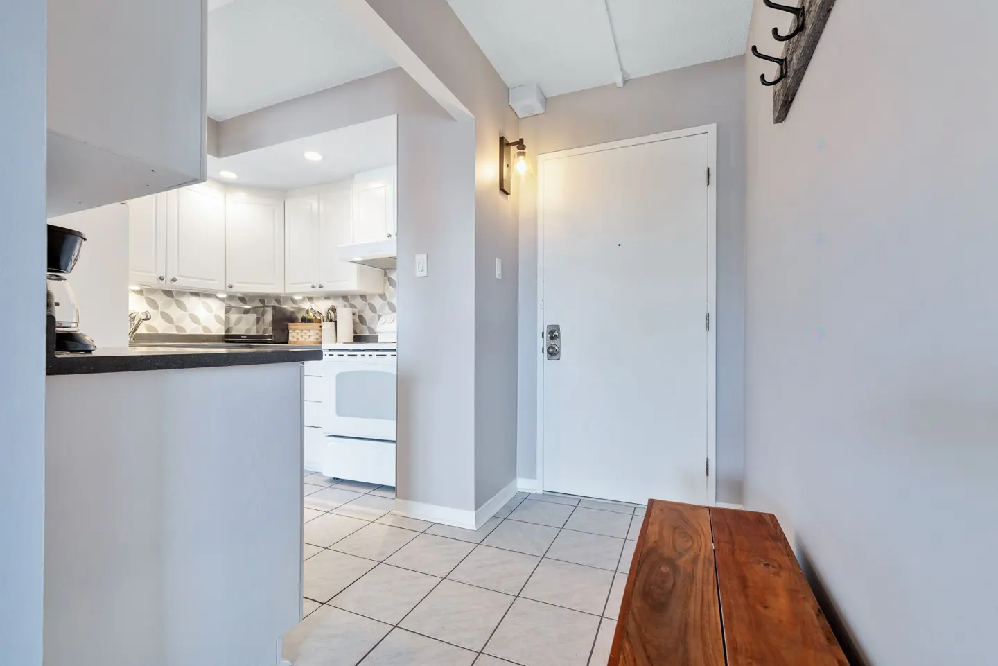 Entryway with a white door, wooden bench, and coat rack. The kitchen is visible with white cabinets and appliances.