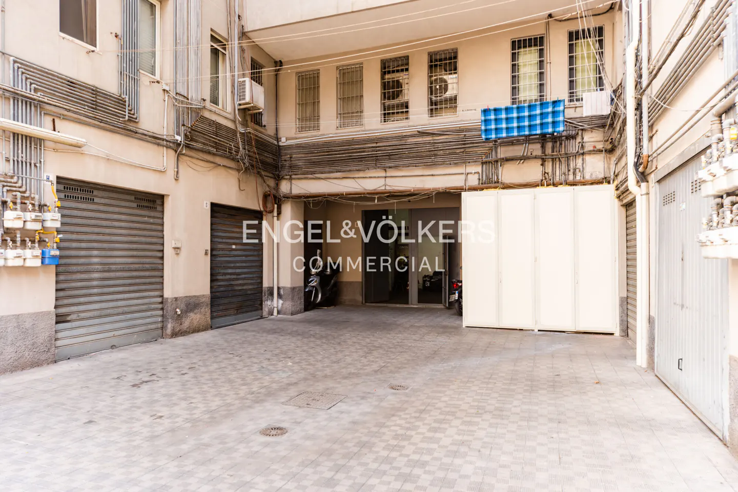Exterior view of a commercial space with gray tile flooring, metal garage doors, and an Engel & Völkers Commercial sign.