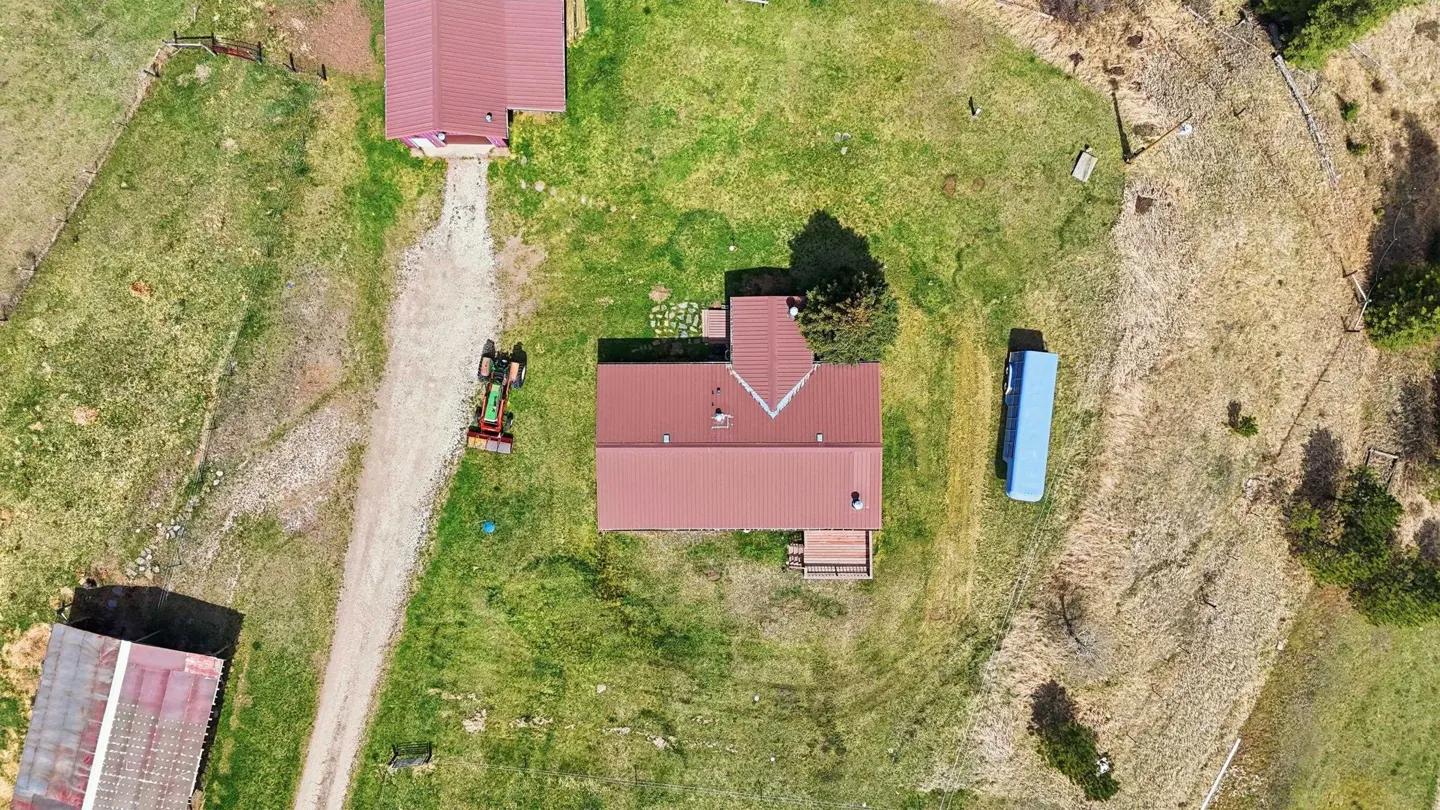 Aerial view of a house with a red metal roof, a tractor on a gravel driveway, and a blue trailer on a grassy rural lot.