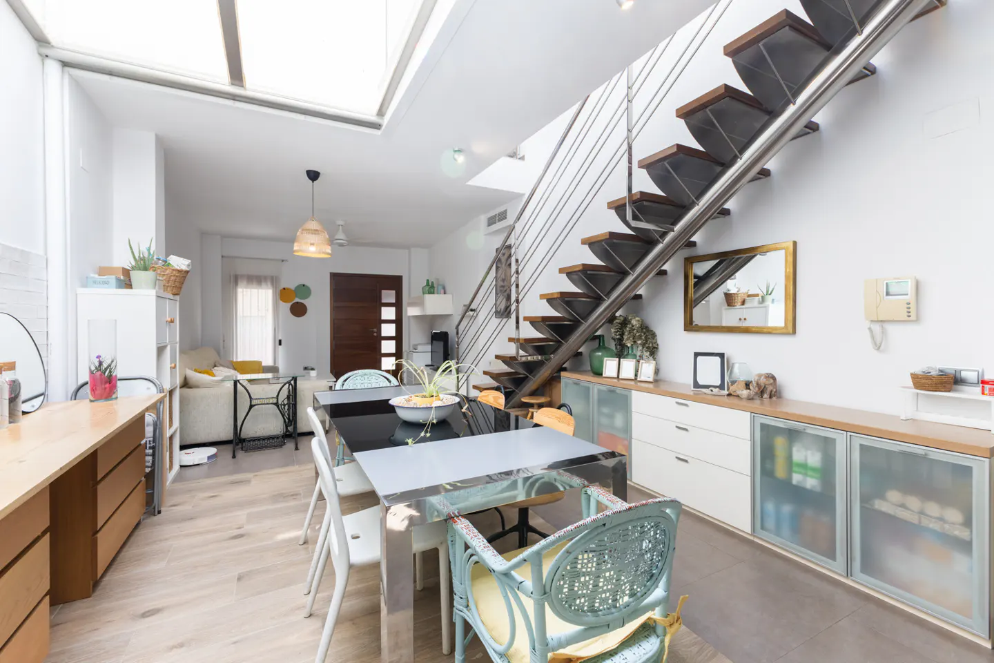 Modern dining area with a glass table, light blue wicker chair, and a floating staircase leading to a bright living room.