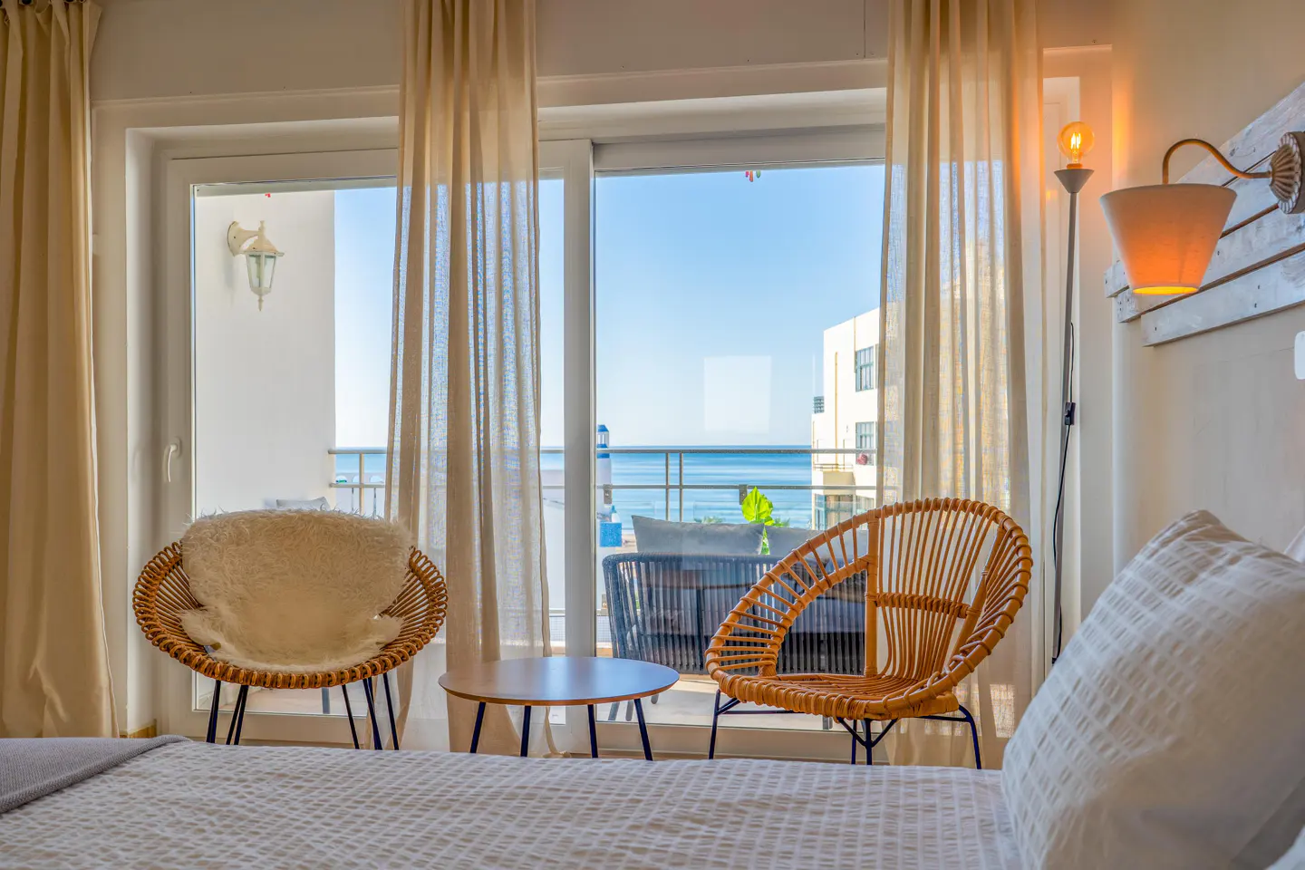 Bedroom with two rattan chairs, a small table, and a balcony view of the ocean through large glass sliding doors.